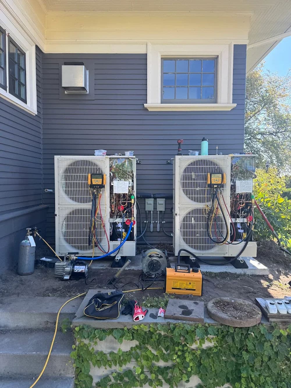 Two outdoor air conditioning units with hoses and tools on a residential home exterior.