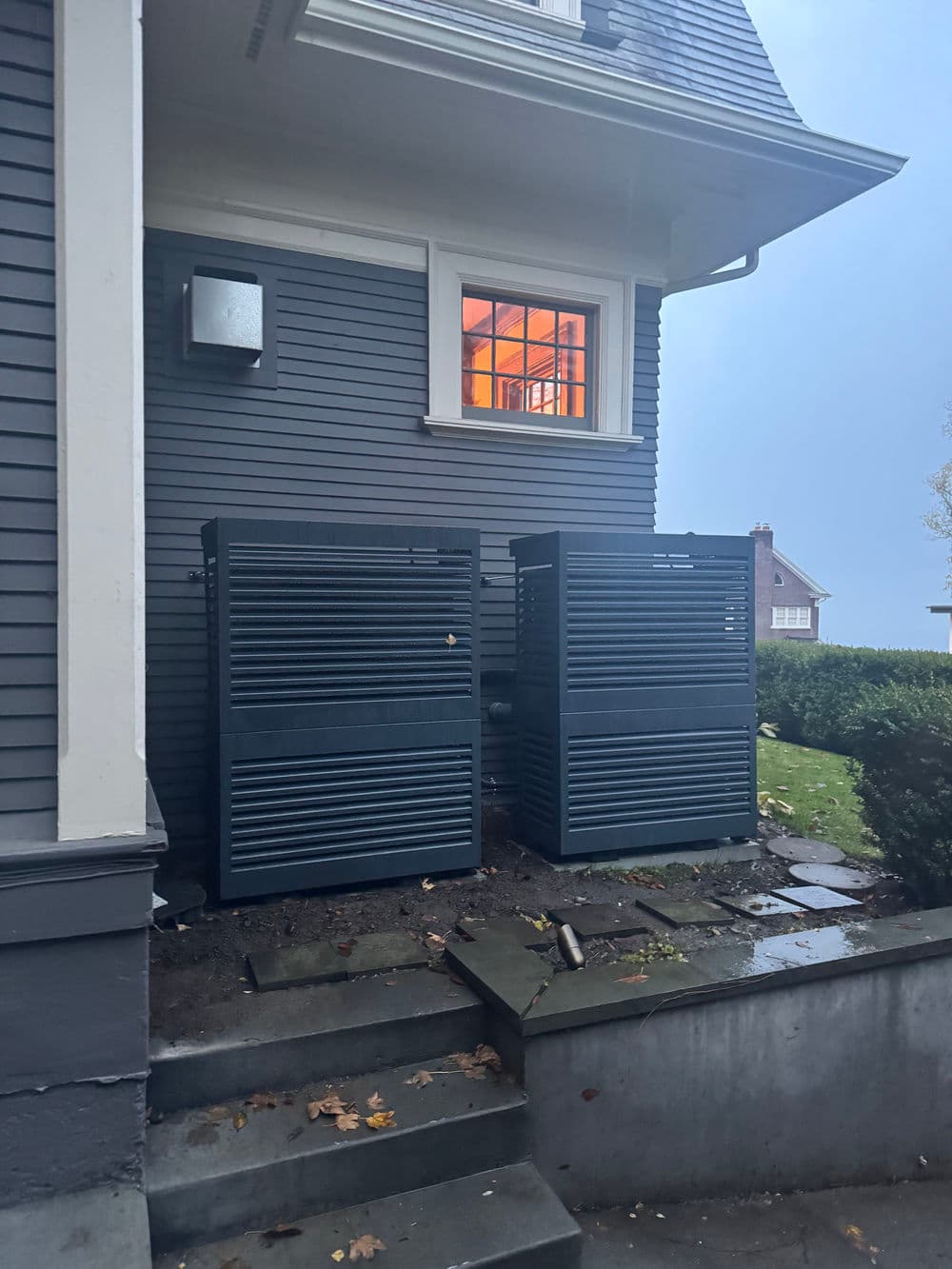 Two dark blue outdoor storage units beside a house with a lit window at dusk.